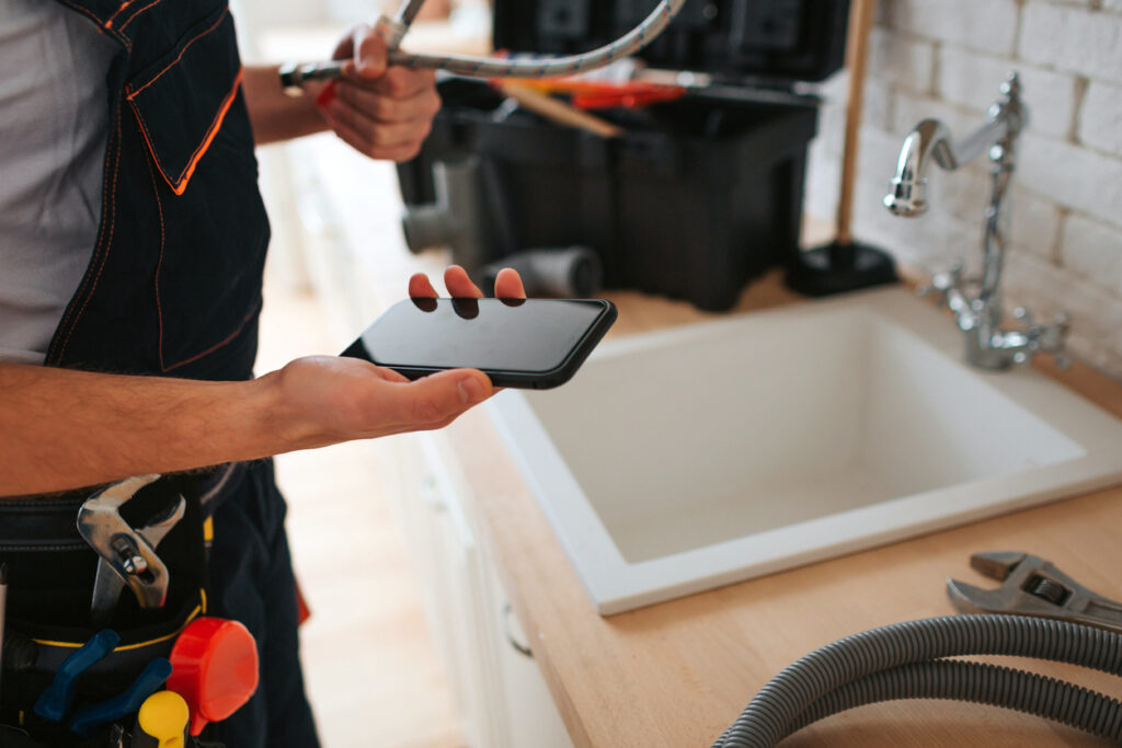 man standing in kitchen at sink, holding a phone and wrench