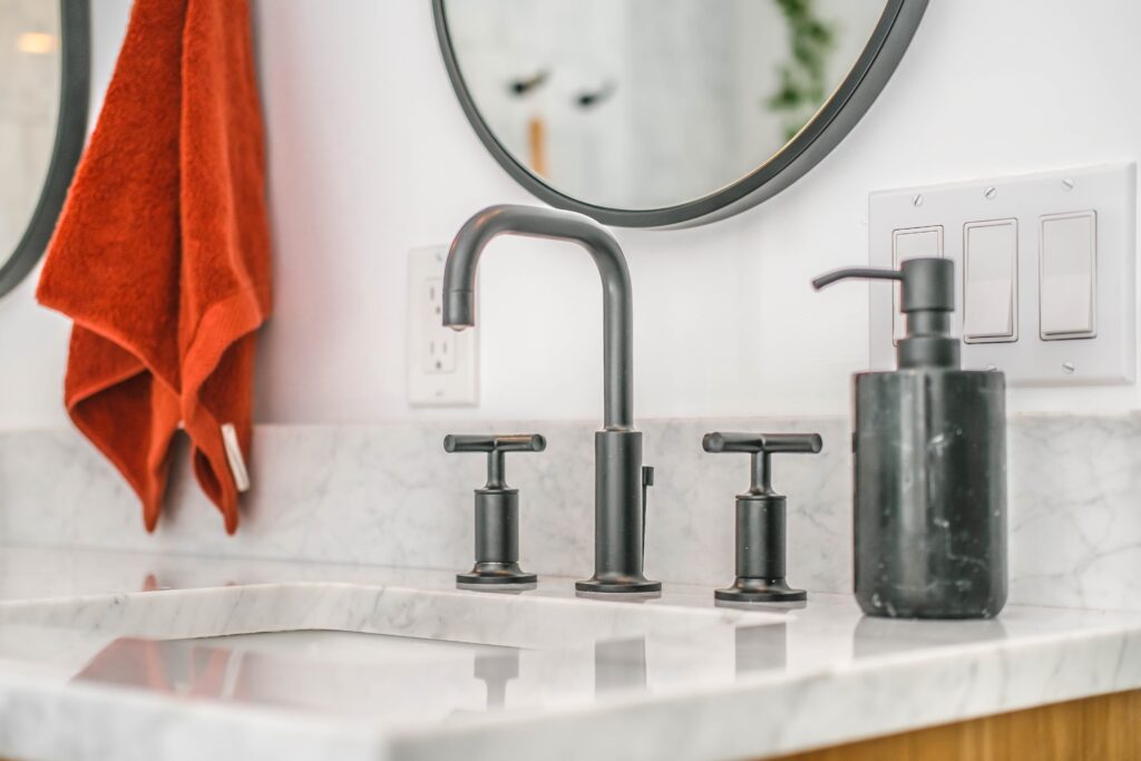 Brushed silver faucet on a granite sink in a bathroom with an orange hand towel, mirror & grey soap dispenser